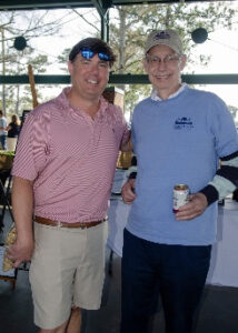 Two men posing together at an outdoor event, smiling; one in a pink polo and khaki shorts, the other in a light blue polo with a cap and a beverage in hand.