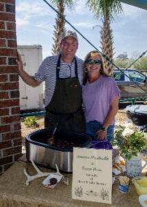 Smiling man in striped shirt and apron and a woman in purple shirt stand beside a large outdoor grill; a handwritten menu sits on the table.