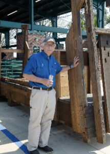 Older man in a blue polo and khaki pants stands beside a timber frame at a construction site, holding a water bottle.