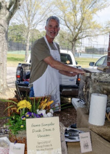 Smiling man in a white apron grills at an outdoor barbecue, with a table of game samples and a handwritten sign nearby.