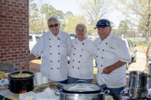 Three chefs in white coats stand behind an outdoor food station, smiling at the camera with pots and slow cookers in front of them.
