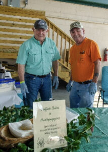 Two smiling men in casual shirts and caps stand behind a table with green plants at an indoor booth exhibition, with a sign describing herbs or a related display behind them.
