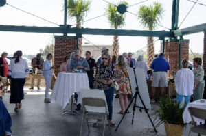 Group of people mingling at an outdoor social gathering under a covered pavilion; a woman in a floral dress stands near a tall white table.