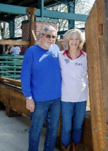 Smiling older couple standing outdoors beside a wooden exhibit structure, posing for the photo.