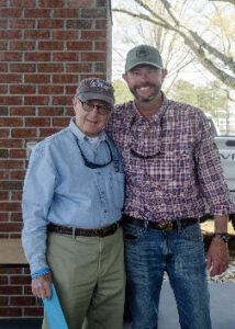 Two men posing together outdoors beside a brick wall, smiling; older man in a light blue shirt and khaki pants, younger man in a red plaid shirt and jeans with caps.