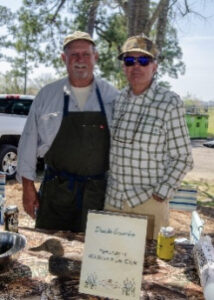 Two men stand behind an outdoor market table with jars and a handwritten sign in front on a sunny day.